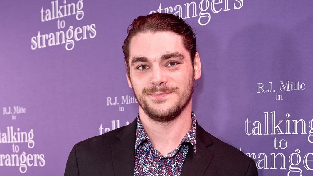 RJ Mitte smiling in front of a purple Talking To Strangers backdrop, wearing a dark jacket and a floral shirt
