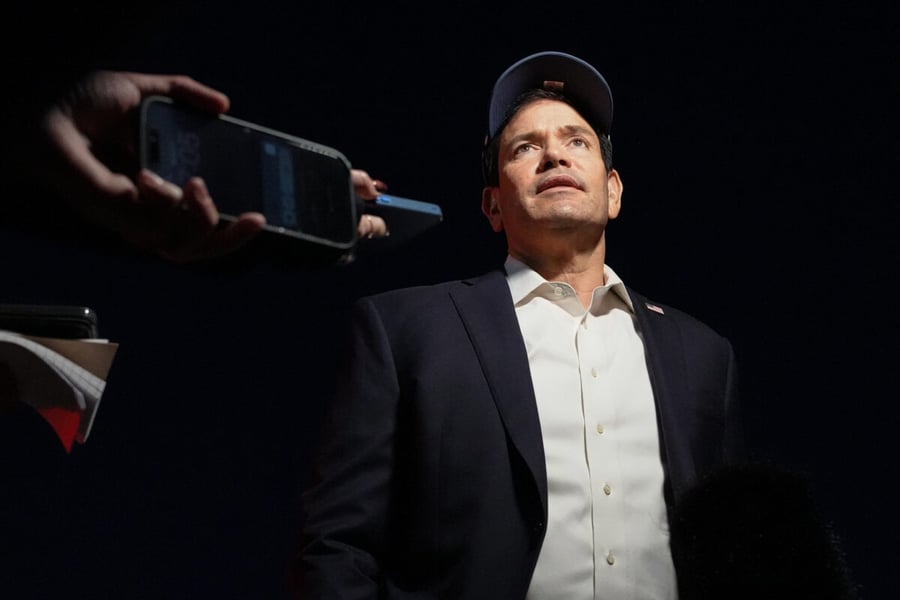 Secretary of State Marco Rubio, in a cap and dark jacket, looks up as reporters hold phones toward him at Joint Base Andrews
