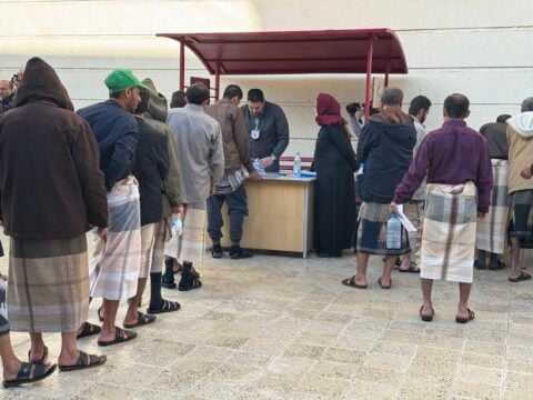 Men in coats and wraparound skirts line up at a desk where a Red Cross staff member processes papers during a detainee release in Sanaa