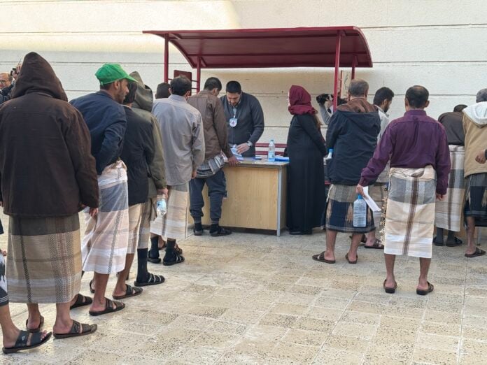 Men in coats and wraparound skirts line up at a desk where a Red Cross staff member processes papers during a detainee release in Sanaa
