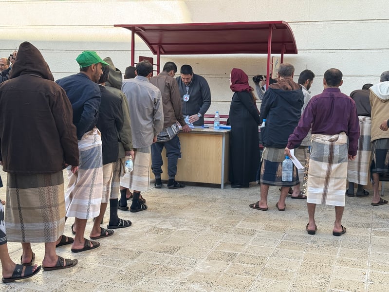 Men in coats and wraparound skirts line up at a desk where a Red Cross staff member processes papers during a detainee release in Sanaa