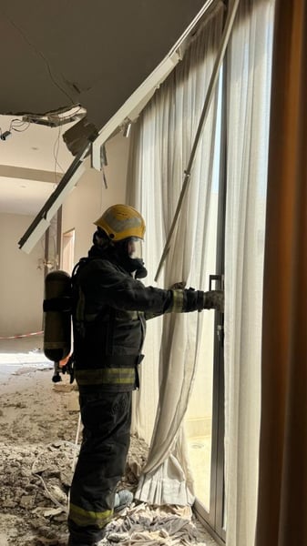 A civil defence worker in a yellow helmet and breathing apparatus inspects a room with a collapsed ceiling and rubble