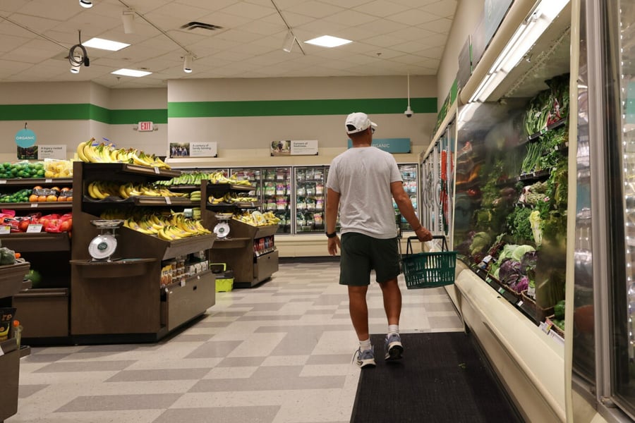 A shopper carrying a basket walks past fruit displays and a refrigerated produce case in a grocery store