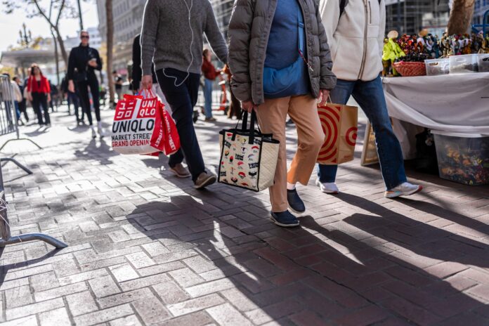 Shoppers carrying retail bags walk along a brick-paved city sidewalk
