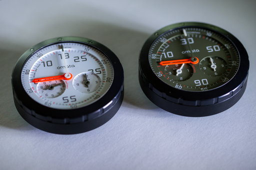 Two Omata cycling computers with light and dark analog dials and orange hands