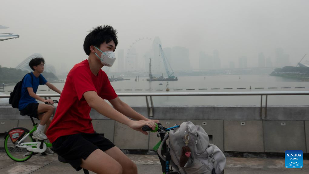 A cyclist in a red shirt and face mask rides past another cyclist on the Marina Bay waterfront, with the Singapore skyline faint in grey haze behind