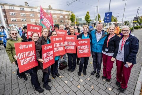 SIPTU members in hospital scrubs and a union official hold red Official Dispute placards beside a tram stop and hospital entrance sign