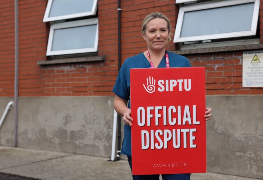 A SIPTU member in blue scrubs holds a red Official Dispute placard in front of a brick building