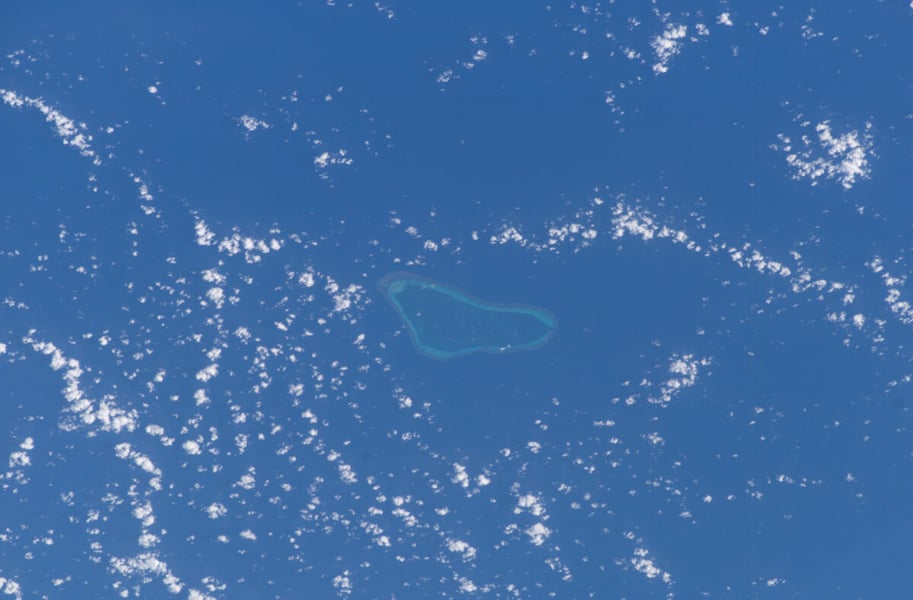 A coral reef with a pale blue lagoon surrounded by deep blue sea and scattered clouds, photographed from space