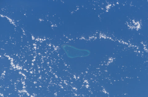 A coral reef with a pale blue lagoon surrounded by deep blue sea and scattered clouds, photographed from space