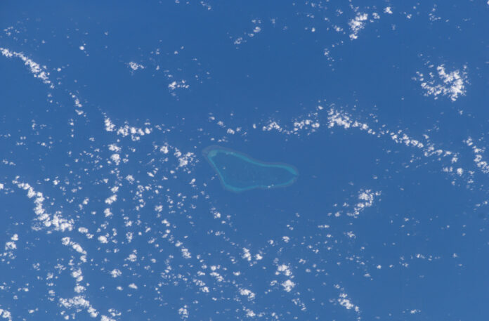 A coral reef with a pale blue lagoon surrounded by deep blue sea and scattered clouds, photographed from space