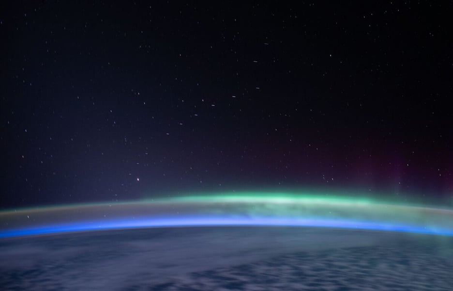 A line of faint streaks, a Starlink satellite train, above a green and blue aurora glowing along Earth's limb