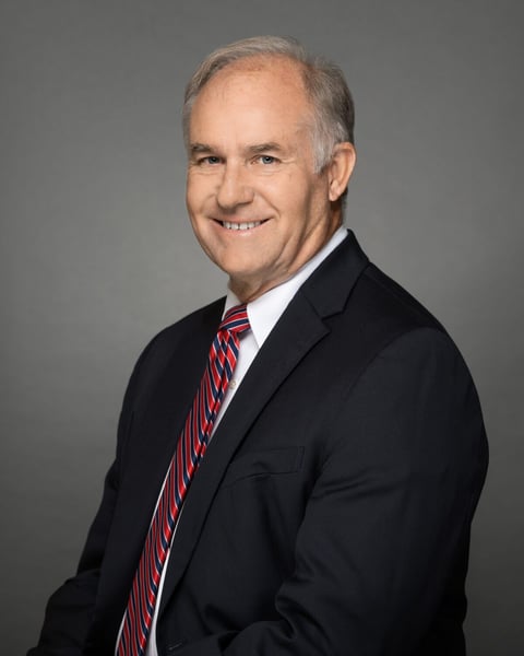 Portrait of Stephen Cash, a smiling man in a dark suit and red-and-blue striped tie against a gray background