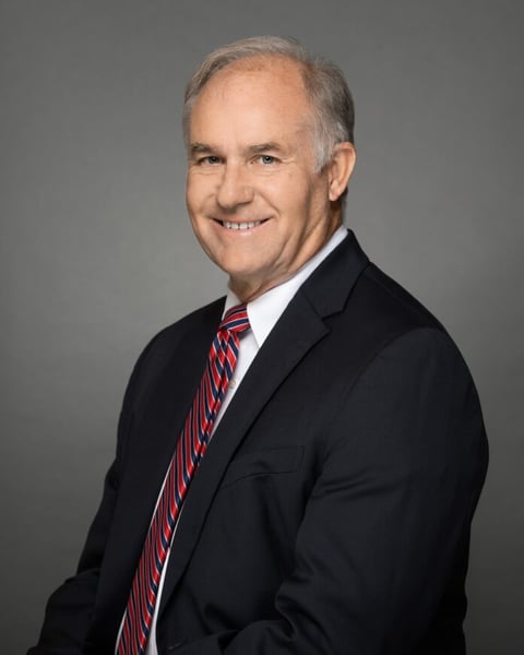 Portrait of Stephen Cash, a smiling man in a dark suit and red-and-blue striped tie against a gray background