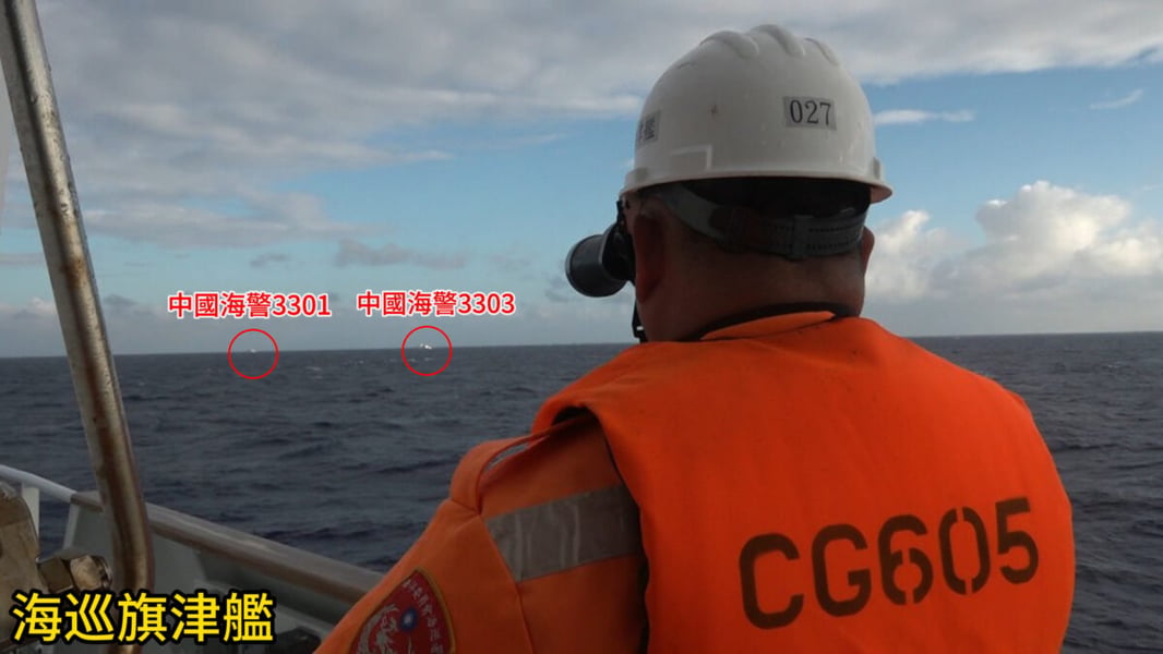 Taiwan Coast Guard crew member in an orange life vest with binoculars watches two distant China Coast Guard vessels, labelled 3301 and 3303, on the horizon