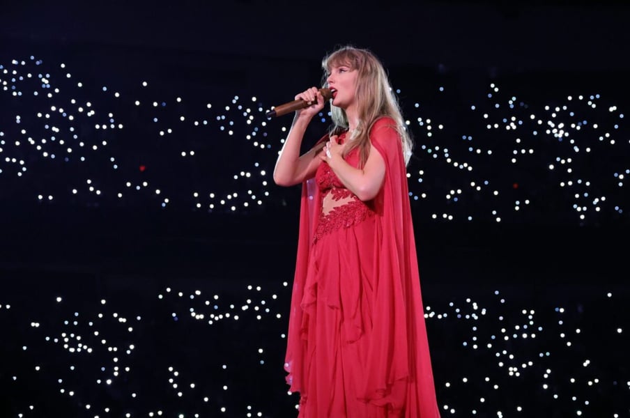 Taylor Swift in a red gown singing into a microphone onstage in front of a field of lights