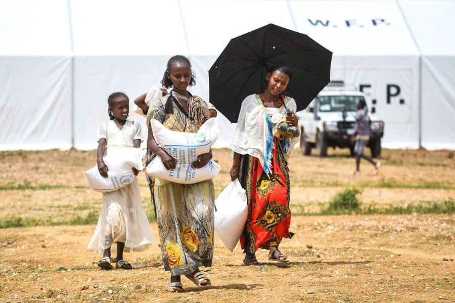 A girl and two women carry sacks of food aid in front of a white WFP storage tent in Tigray, Ethiopia