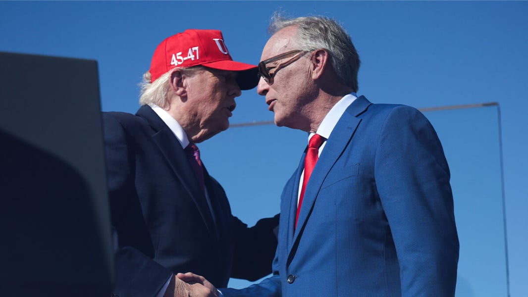 President Trump in a red cap and Rep. Andy Biggs in sunglasses and a blue suit shake hands on stage under a blue sky