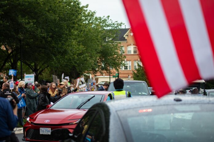 Demonstrators with signs outside Trump National Golf Club as cars arrive for the first meme coin dinner