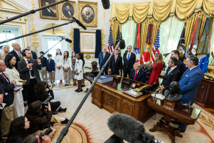 President Donald Trump seated at the Resolute Desk in the Oval Office during an announcement on Trump Accounts, with reporters and photographers in the foreground
