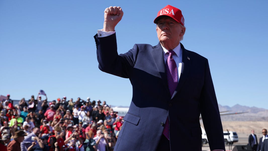 President Trump in a red USA cap raises a clenched fist in front of a crowd at an outdoor rally