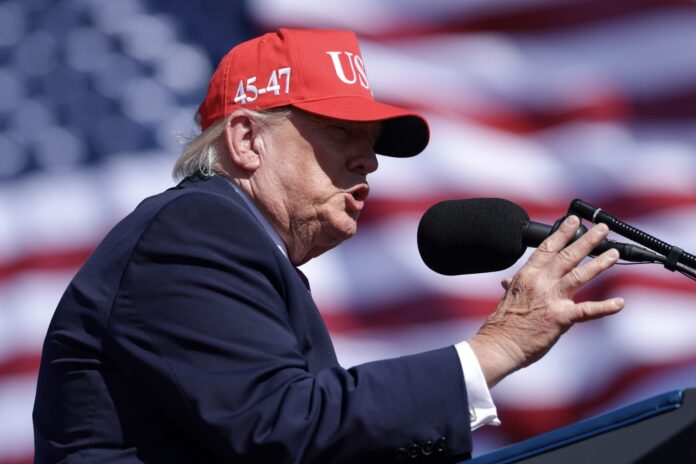 President Donald Trump in a red cap speaks into a microphone in front of an American flag at a rally in Bullhead City, Arizona