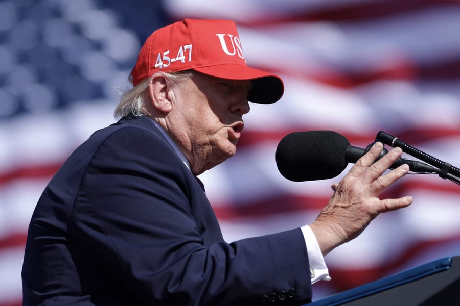 President Donald Trump in a red cap speaks into a microphone in front of an American flag at a rally in Bullhead City, Arizona