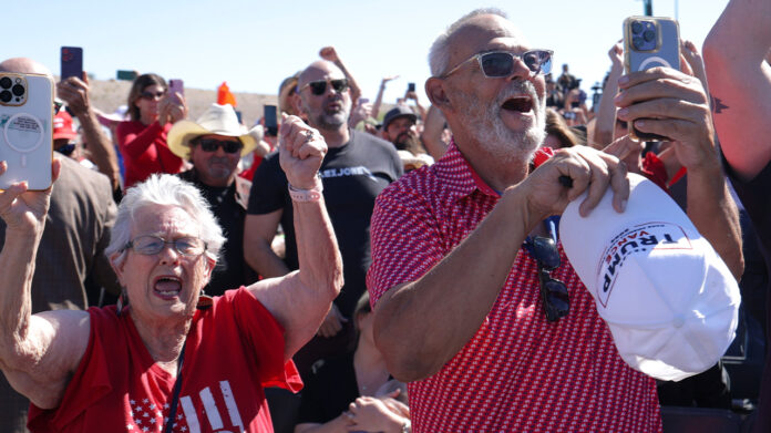 Supporters in red shirts and Trump caps cheer and film on phones at an outdoor rally