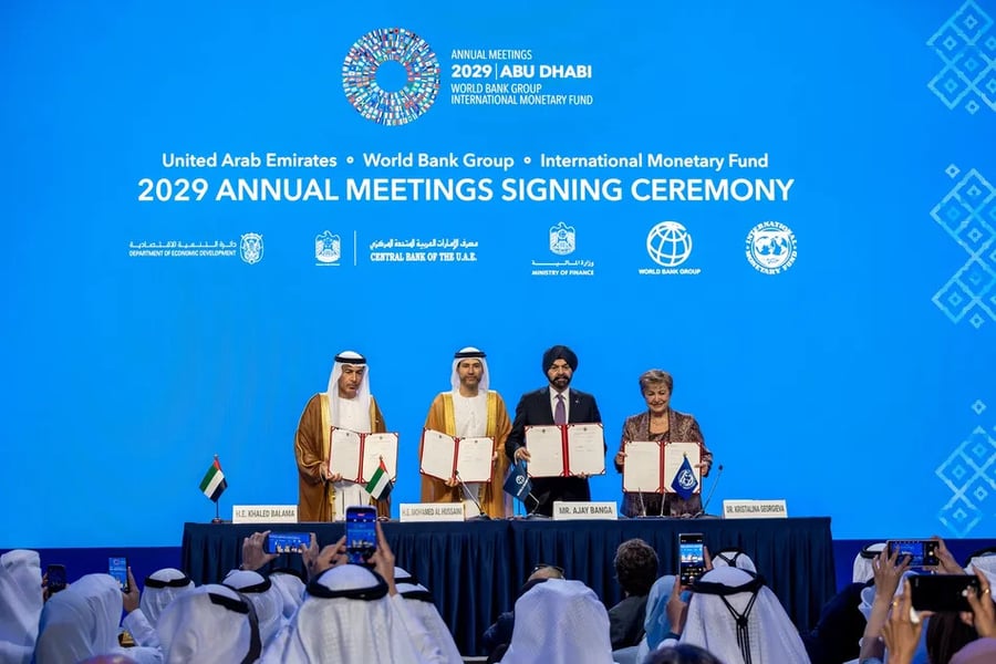 Four signatories stand behind a table holding signed documents under a 2029 Annual Meetings Abu Dhabi signing ceremony banner while the audience films on phones