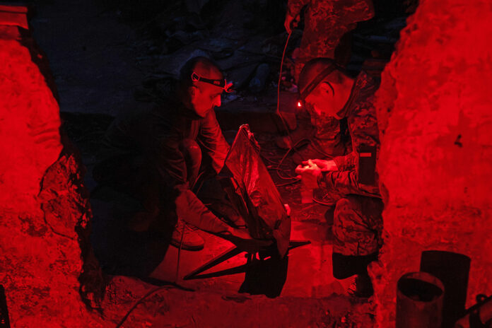 Two Ukrainian soldiers with head torches crouch in a trench lit red as one handles a Starlink dish