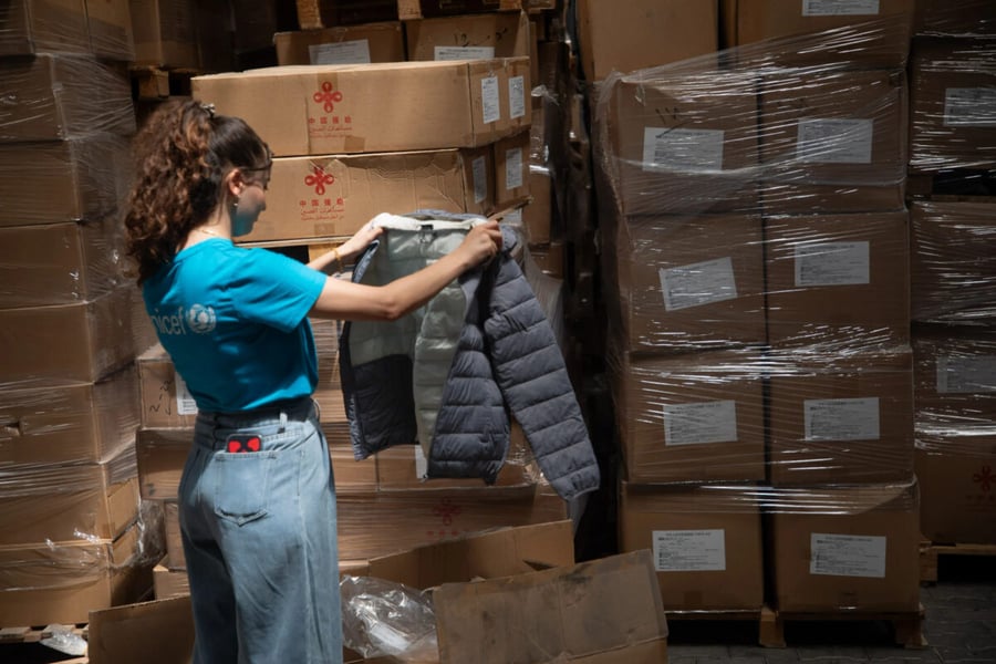 A UNICEF staff member in a blue T-shirt holds up a grey padded children's jacket among stacked, shrink-wrapped boxes in a warehouse