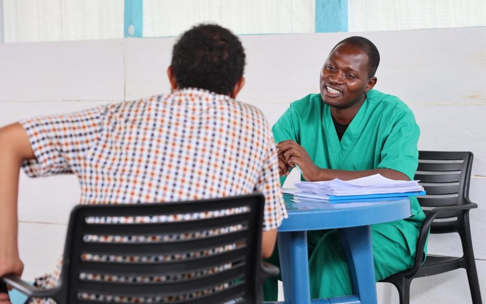 A health worker in green scrubs sits across a blue table from a person with their back to the camera during a counselling session in Bunia