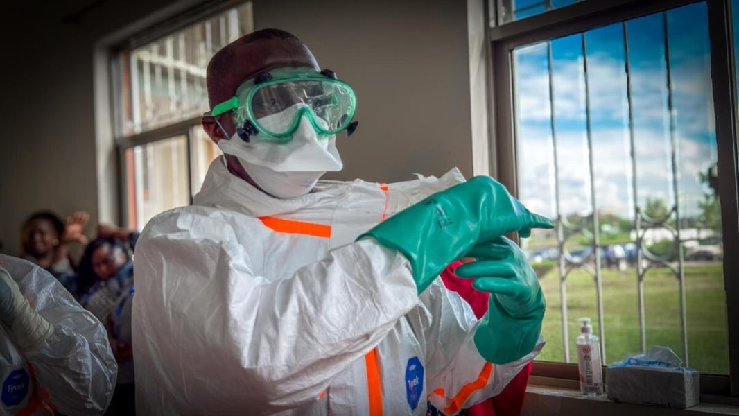 Health worker in a white suit, goggles and mask pulling on green gloves beside a window