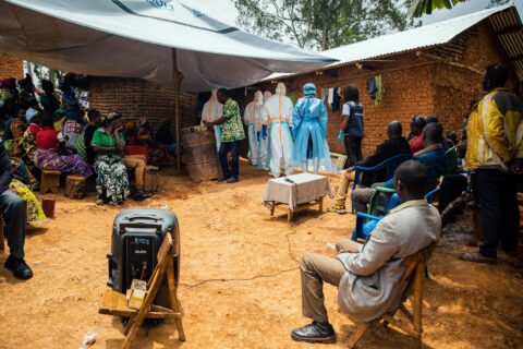 Responders in white protective suits stand under a tarpaulin among seated mourners at a funeral gathering in Ituri Province, Congo