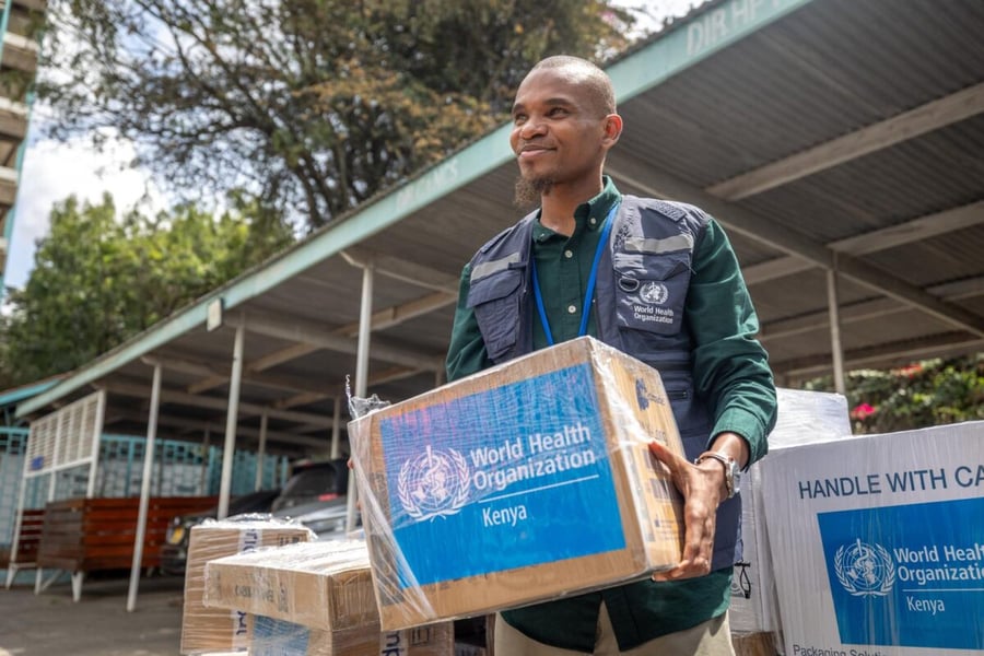 A smiling man in a vest carries a cardboard box marked World Health Organization Kenya beside stacked supply boxes