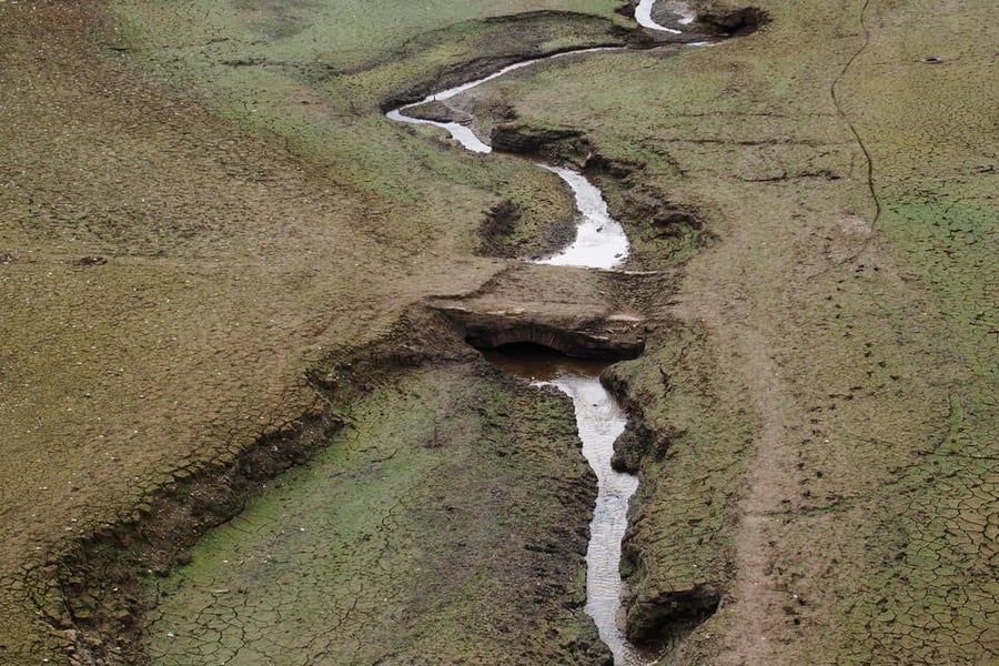 Cracked reservoir bed at Wimbleball with a stream winding past the stone arch of Bessom Bridge, exposed as water levels have dropped