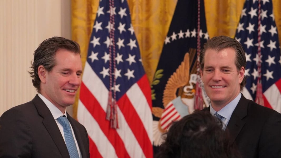 Cameron and Tyler Winklevoss smiling in front of American flags at the White House
