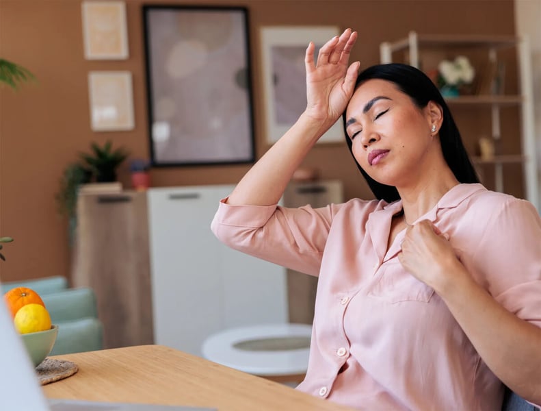 Woman in a pink blouse closing her eyes and touching her forehead during a hot flash at a table in her home