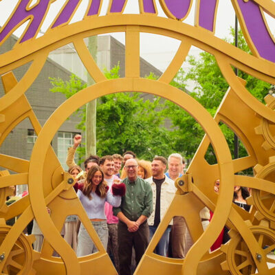Contestants and onlookers smile behind a large golden gate of interlocking gears in a promotional still from Netflix's Wonka's The Golden Ticket