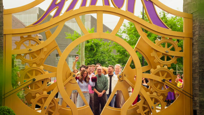 Contestants and onlookers smile behind a large golden gate of interlocking gears in a promotional still from Netflix's Wonka's The Golden Ticket