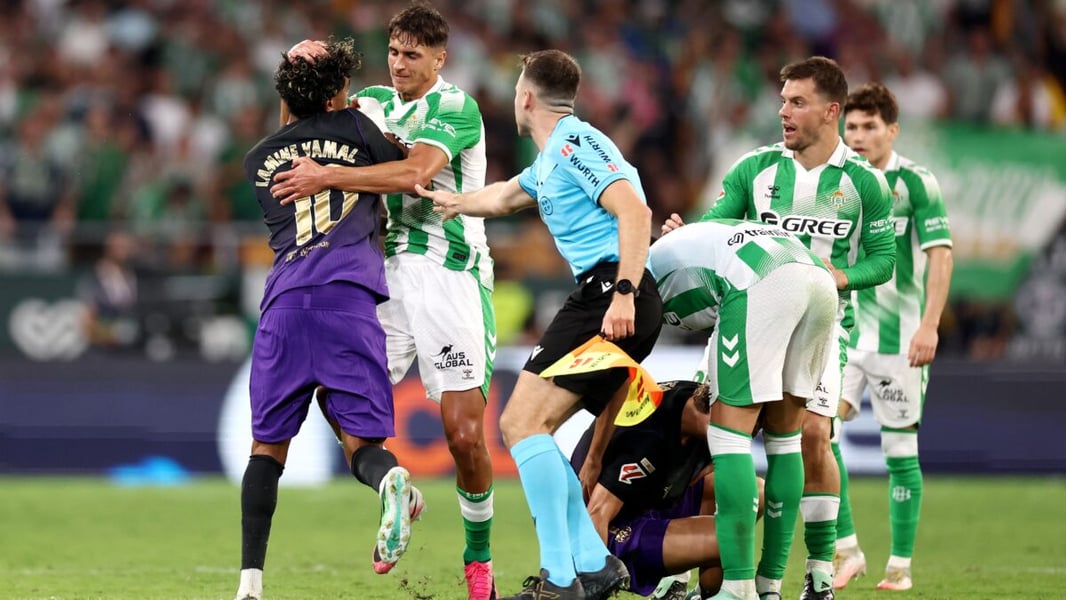 Lamine Yamal in Barcelona's purple kit is held by a Real Betis player as a match official steps between them