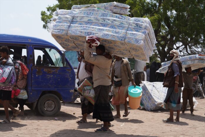 Men carry stacked mattresses on their heads beside a blue minibus as displaced families receive supplies in Aden, Yemen