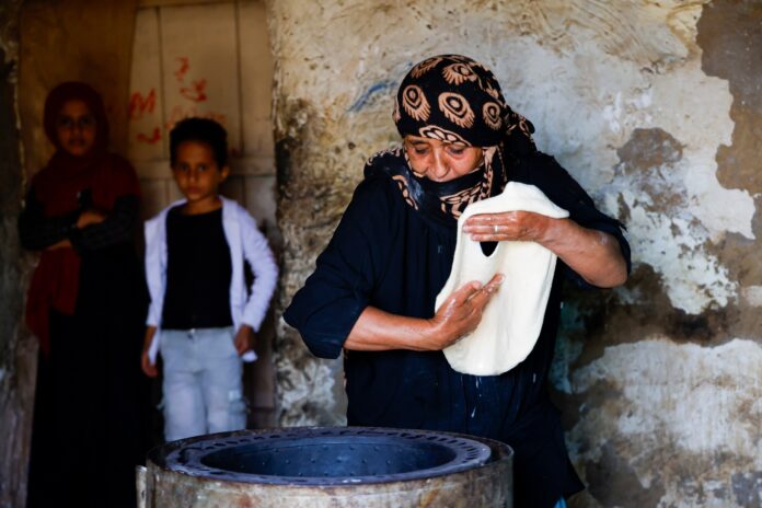 A woman in a black headscarf shapes dough beside a clay oven while two children watch