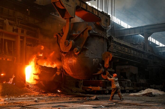A steelworker in a hard hat guides a ladle hook beside a glowing furnace opening in the open-hearth shop at Zaporizhstal