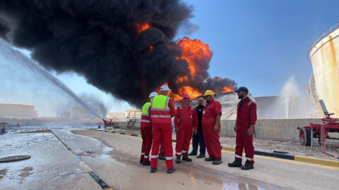 Workers in red overalls and hard hats stand on a wet road in front of a burning fuel tank and a large plume of black smoke at an oil refinery