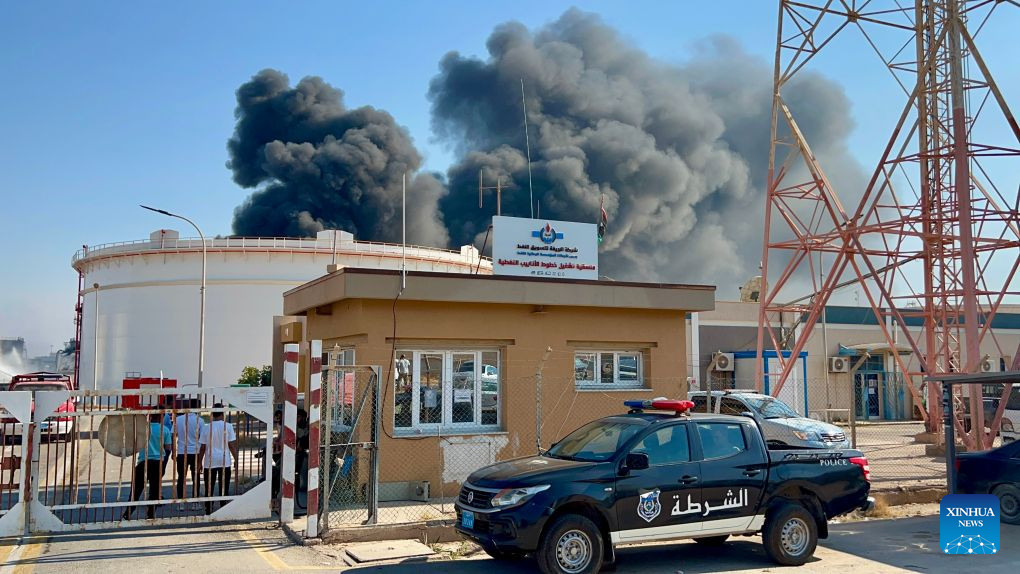 A black police pickup truck parked by a guard post with a white storage tank and a dark plume of smoke rising behind it