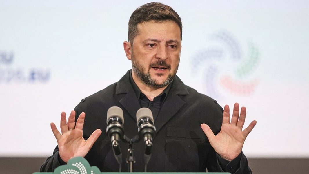 Volodymyr Zelenskyy gestures with both hands while speaking at a podium with two microphones